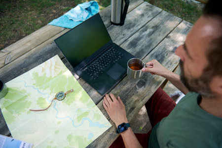 Man sitting at outdoor table with laptop, map, and cup of tea. Concept of planning, adventure, and...