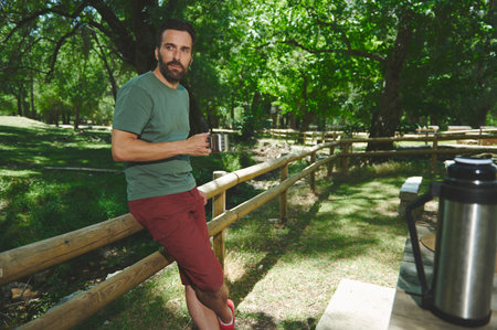 Outdoor scene with a relaxed man holding a coffee mug, standing near a wooded area with a picnic setup, evoking a sense of calm and natural enjoyment.の写真素材