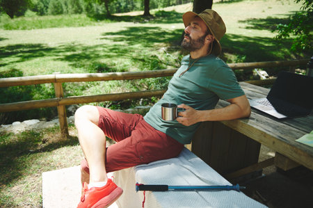 A relaxed man with a cup sits under the shade by a table with a laptop during a sunny day in a serene natural setting. A trekking pole lies on the spot.の写真素材