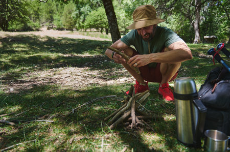 A man arranges sticks to start a campfire during a camping trip in a lush forest. He is surrounded by camping gear in the sunny wilderness, embodying the spirit of outdoor adventure and nature.の写真素材