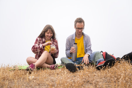 A pair of friends enjoying a relaxing moment outdoors, having a light meal and beverages while seated on a dry grassy hill under a cloudy sky, emphasizing adventure and togetherness.の写真素材