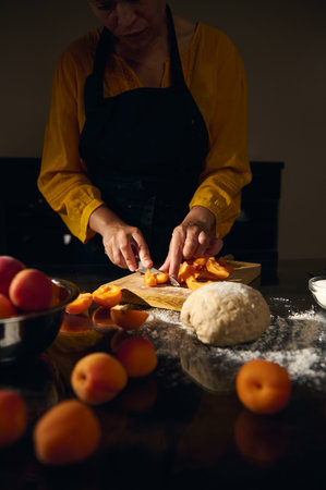 A woman in a yellow blouse chops fresh apricots on a wooden board. Flour and dough are prepared for...