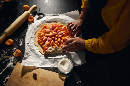 A close-up view of a person shaping a galette with fresh apricots, showcasing a warm ambiance and the art of homemade baking. Spices and utensils enrich the scene.の写真素材