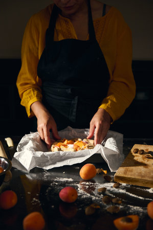 A woman wearing a black apron arranges fresh fruit and baking ingredients on parchment paper in a...