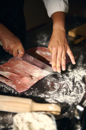 A chef works with pink pasta dough, meticulously cutting it on a floured workspace, showcasing...