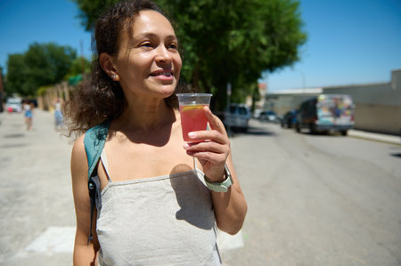 A cheerful woman carrying a backpack and holding a cup with a fruity beverage enjoys a bright summer...