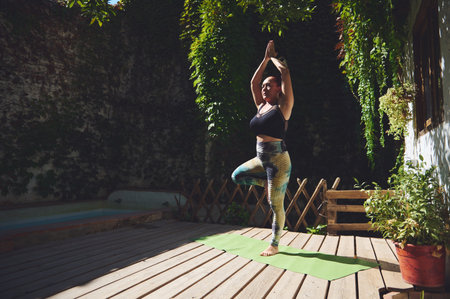 A young woman skillfully performing the tree yoga pose on a wooden deck surrounded by lush green foliage, illustrating harmony and mindfulness in an outdoor wellness setting.の写真素材