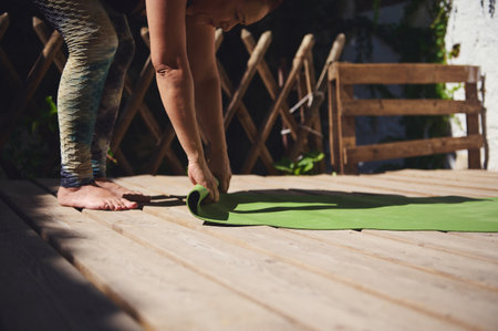 A person unrolling a green yoga mat on a wooden deck in a peaceful outdoor atmosphere surrounded by greenery, suggesting preparation for relaxation or exercise activities in open air.の写真素材