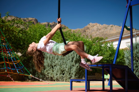 A happy young girl enjoying the day as she swings on a rope at a vibrant playground under a bright...
