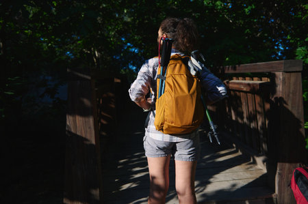 A person hikes on a forest trail, crossing a wooden bridge under sunlight. They carry a yellow...