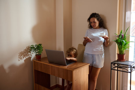 A mother enjoys a warm drink while managing her remote work and child in a cozy, sunlight-lit living space with plants.の写真素材