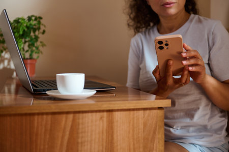 A woman engages with her smartphone at a wooden desk, featuring a laptop, coffee cup, and a...