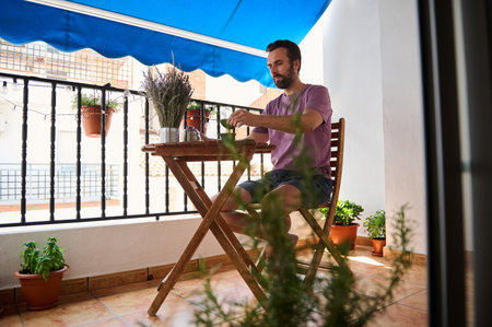 A serene outdoor scene of a man sitting at a wooden table, enjoying tea on a balcony decorated with potted plants and under a blue shade awning.の写真素材