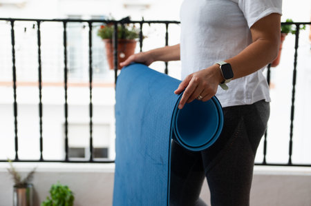 A woman rolling a blue yoga mat on a balcony, wearing athletic attire, representing fitness and mindfulness. The relaxed setting highlights the importance of outdoor environments for calm exercises.の写真素材