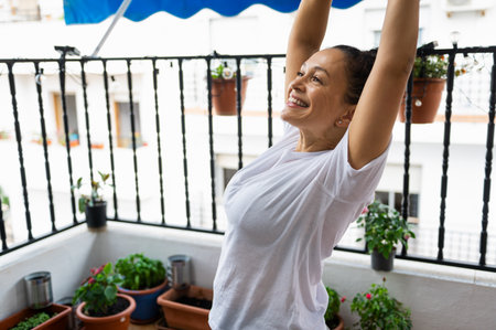 A woman standing on her balcony, smiling brightly while stretching her arms upwards. Around her are vibrant potted plants and a peaceful outdoor setting, evoking feelings of joy and tranquility.の写真素材