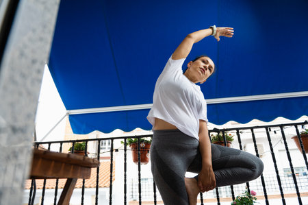 A woman stretches in a yoga position on a balcony, enjoying outdoor fitness under a bright blue...