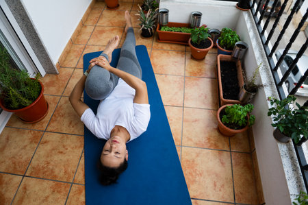 A woman performs a yoga pose on a blue mat located on an apartment balcony with various potted plants and greenery. The scene captures relaxation, healthy living, and the tranquility of home fitness.の写真素材