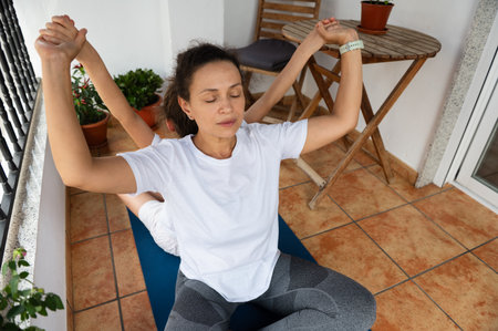 A woman sitting cross-legged practicing yoga with her child on a sunny balcony. The scene...