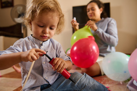 A young child concentrates on inflating a balloon with a manual pump, assisted by a cheerful parent in a cozy home setting. The image conveys teamwork, family bonding, and joy.の写真素材