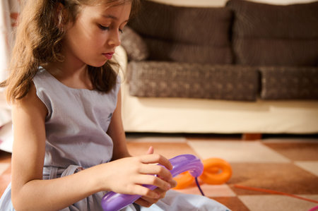 A young girl carefully twists a purple balloon while sitting indoors. The warm, cozy home background and her focused expression highlight her creativity and concentration during this playful activity.の写真素材