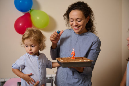 A mother holds a cake with a lit candle for her child's second birthday celebration, surrounded by...