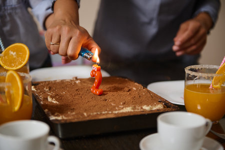 A close-up of hands lighting a number two candle on a cake, surrounded by beverages and cups. The scene showcases a celebratory atmosphere with attention to detail and cozy emotions.の写真素材