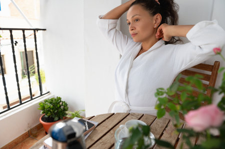 A serene morning scene featuring a woman enjoying relaxation in a white bathrobe on a balcony surrounded by flowers and coffee.の写真素材