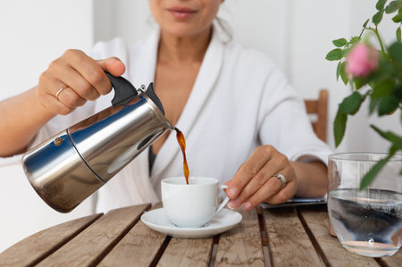 A serene moment enjoying fresh coffee poured from a stovetop kettle into a white ceramic cup outdoors. The setting features a wooden table adorned with some flowers.の写真素材