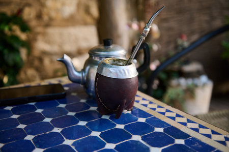 A mate cup with bombilla straw and a teapot on a decorative blue tiled table. The scene portrays a calm atmosphere, suggesting leisure and tradition, perfect for culturally-inspired visuals.の写真素材