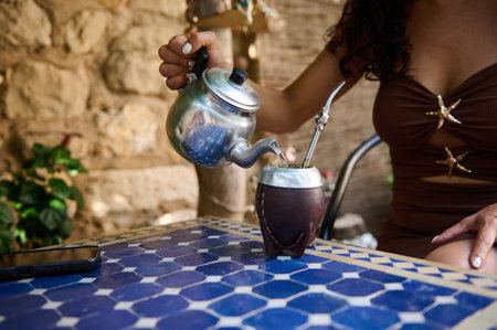 A woman seated outdoors pours tea into a traditional cup on a patterned blue table, creating a...