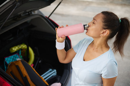 Young woman in casual fitness attire drinking from a pink water bottle near her open car trunk....