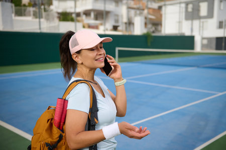 A woman wearing sports attire and a cap is standing on a tennis court, smiling while engaged in a phone call. She carries a backpack and has tennis rackets, suggesting a fitness-oriented activity.の写真素材