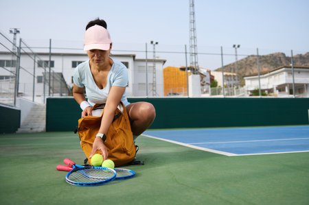 A sportswoman organizes her equipment, including tennis rackets and balls, at a tennis court. She appears focused and active. The modern setting underlines her dedication to staying fit.の写真素材