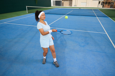 A young woman on an outdoor tennis court practicing her swing. Wearing casual sportswear, she demonstrates athletic commitment amid a vibrant blue court. Capturing determination and enthusiasm.の写真素材