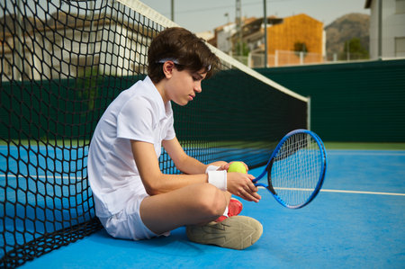 A young tennis athlete in white clothing sits on a vibrant blue tennis court, resting by the net while holding a racket and tennis balls. The composition emphasizes sports and determination.の写真素材