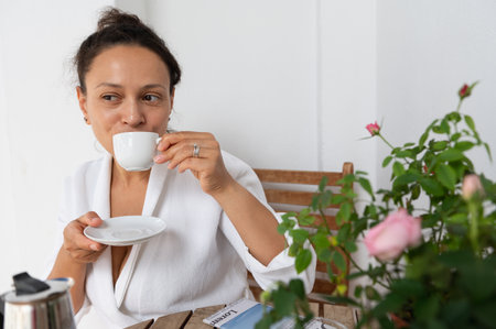 A woman enjoys her morning coffee on a patio, surrounded by flowers and a serene ambiance. The table features a coffee pot, cup, and saucer, reflecting a peaceful morning setting.の写真素材