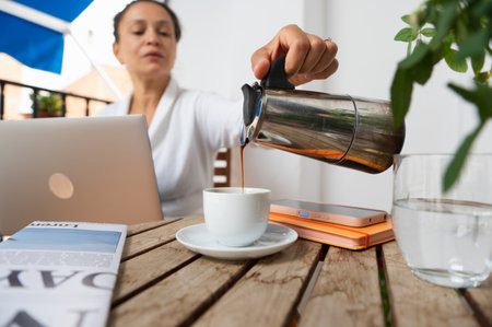 A woman works on her laptop, enjoying coffee while seated at a wooden table outdoors. The image conveys a serene and productive atmosphere for remote work or leisure.の写真素材
