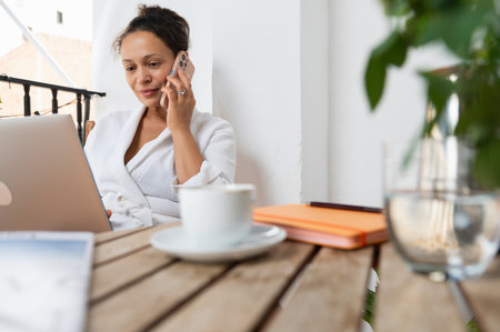 A woman multitasking in a serene setting, engaging in a phone conversation while working on her laptop. The image symbolizes remote work, productivity, and communication in a relaxed environment.の写真素材