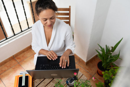 A focused woman types on a laptop while sitting on a stylish balcony. Surrounded by potted plants and natural light, she engages in remote work, enjoying a productive and relaxed atmosphere.の写真素材