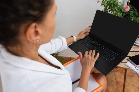 A woman works on her laptop outdoors, seated at a table with a nearby notebook and plants.の写真素材