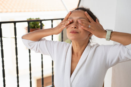 A serene woman stands on a balcony surrounded by soft outdoor light wearing a white bathrobe.の写真素材