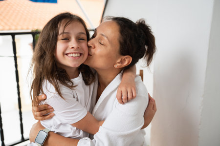 A mother warmly kisses her cheerful, smiling daughter while hugging her affectionately indoors.の写真素材