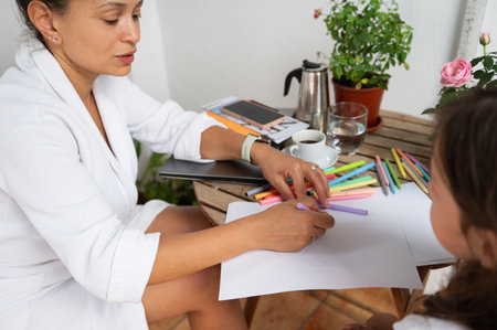 A woman and a child sit at a wooden table outdoors, engaging in a creative drawing activity with colorful pens. The cozy setting features plants and a cup of coffee, emphasizing relaxation and connection.の写真素材