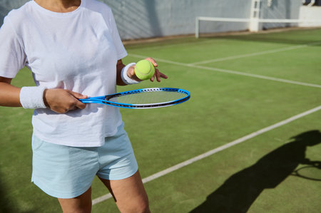 Close-up of a tennis player holding a racket and ball on a sunny outdoor court, ready to play. The green court and white lines are visible, reflecting an active sports environment and preparation.の写真素材