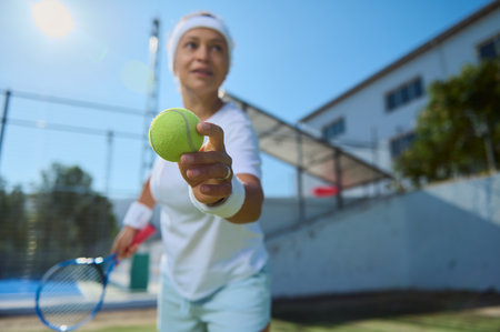 Focused woman holding a tennis ball, mid-action during a game on a bright, sunny day. A tennis court stretches behind her, emphasizing the outdoor and athletic setting.の写真素材