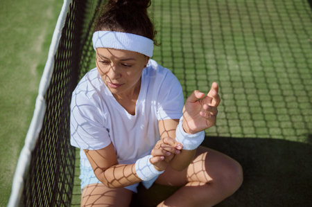 A young woman wearing sports attire takes a break by sitting on the tennis court, with sunlight casting shadows through the net. Her relaxed posture conveys a blend of determination and calm.の写真素材