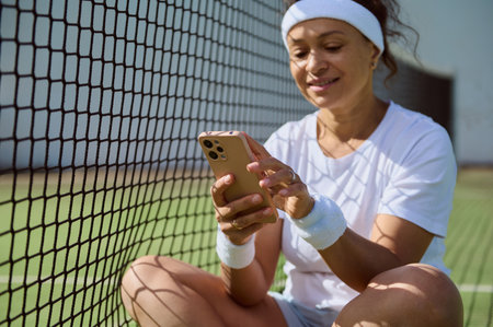 A woman sits cross-legged by a tennis net, smiling and using her smartphone. She appears relaxed, wearing tennis attire, including a white headband and wristbands. The scene suggests leisure and connectivity.の写真素材