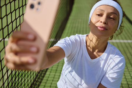 A stylish woman wearing a headband and tennis attire takes a selfie on a sunny tennis court. Emphasizing leisure and sports, her smiling expression captures the joy of an outdoor activity.の写真素材