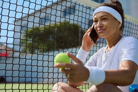 A cheerful woman in sports attire sits on a tennis court, talking on the phone while holding a tennis ball, highlighting a balance between fitness and communication in a relaxed, outdoor setting.の写真素材