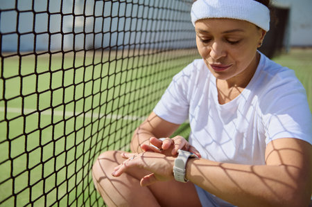 A woman wearing athletic gear rests by a tennis net while interacting with her fitness device. The image captures relaxation, focus, and a healthy outdoor setting on a sunny day.の写真素材